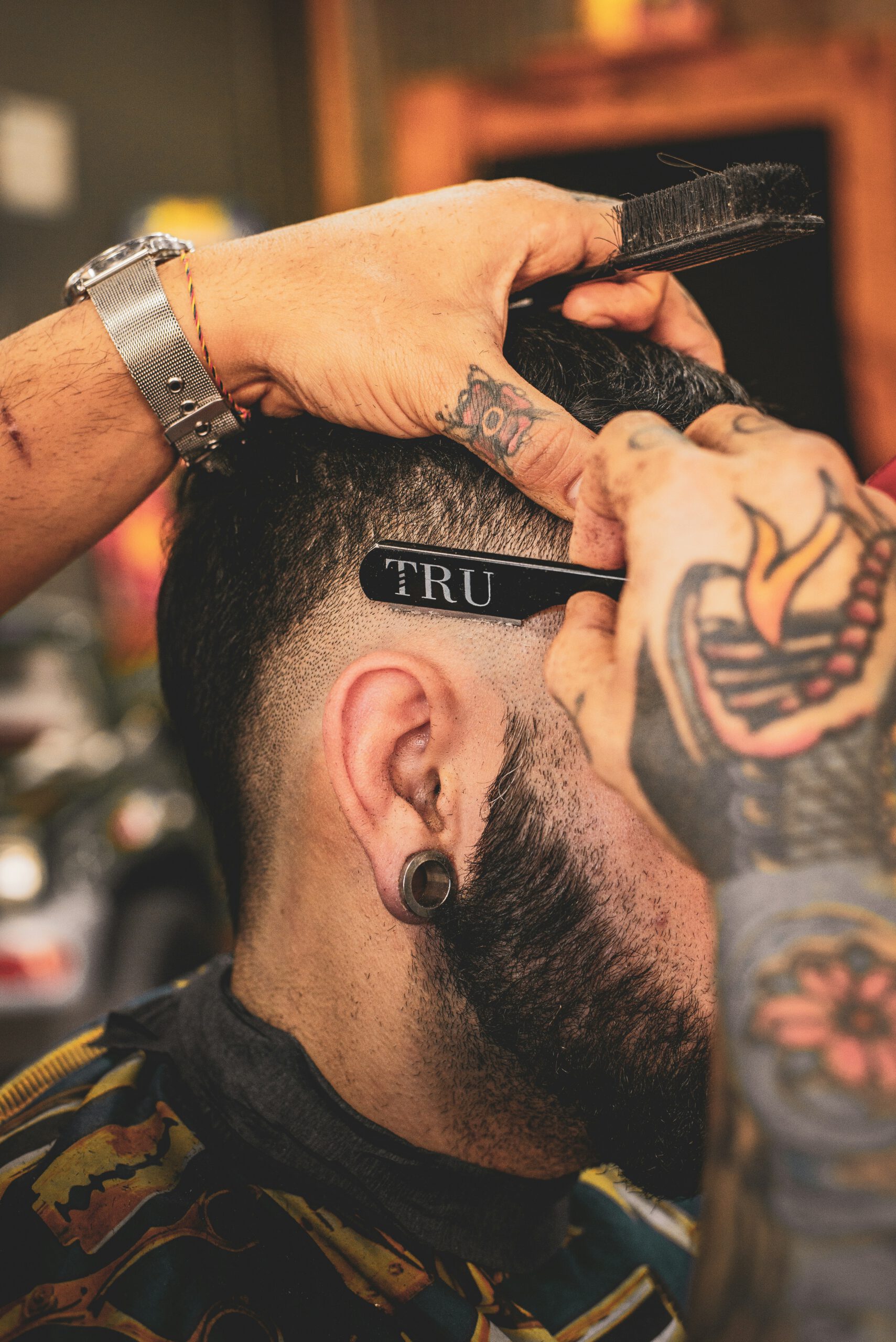 Un barbier aux bras tatoués dessine la racine des cheveux d'un homme à l'aide d'un rasoir droit portant l'inscription 