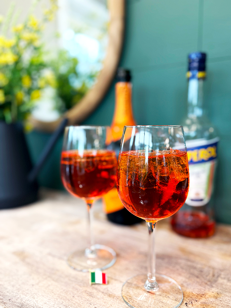 Deux verres de cocktails spritz à l'orange avec de la glace sur une surface en bois, avec Aperol et une autre bouteille floue à l'arrière-plan. Un petit cure-dent représentant le drapeau italien se trouve au premier plan, ainsi que des fleurs dans un vase.